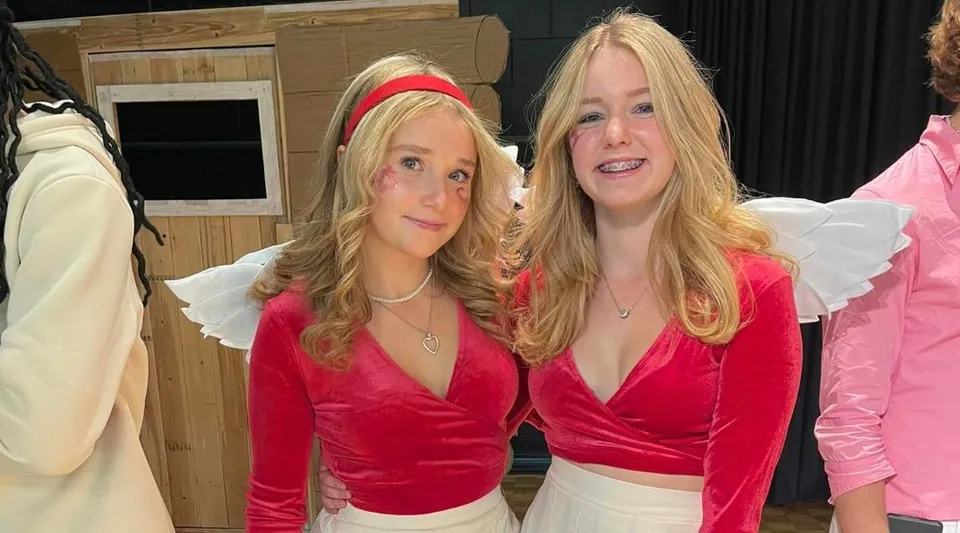Due studentesse del Vankleek Hill Collegiate Institute indossano maglie di velluto rosso e stanno in piedi davanti a stender per abiti e a una struttura in legno.