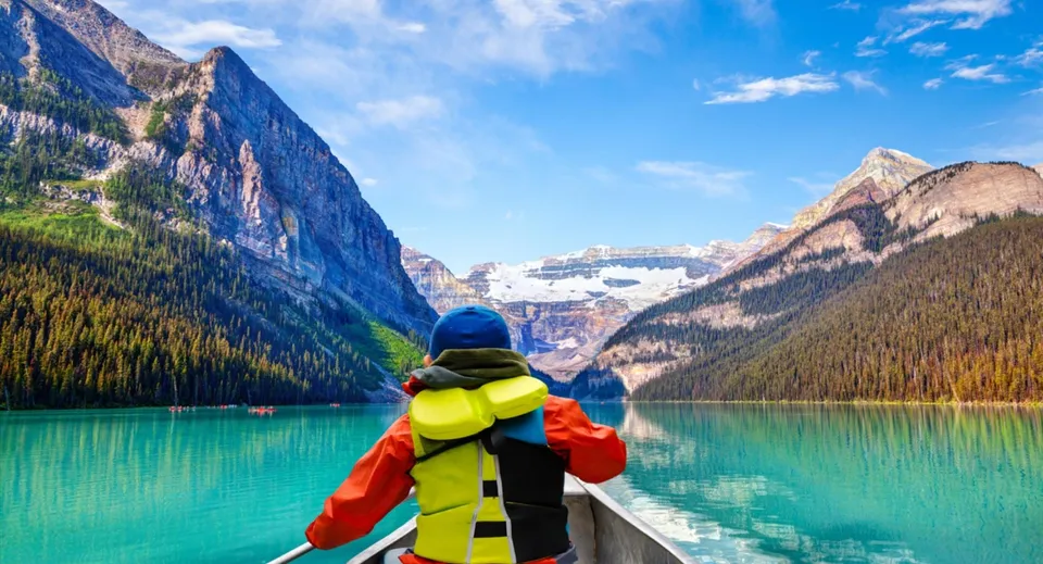 Una persona con una colorata attrezzatura outdoor sta in piedi su una barca su un lago davanti a montagne innevate presso la Clayton Heights Secondary School.