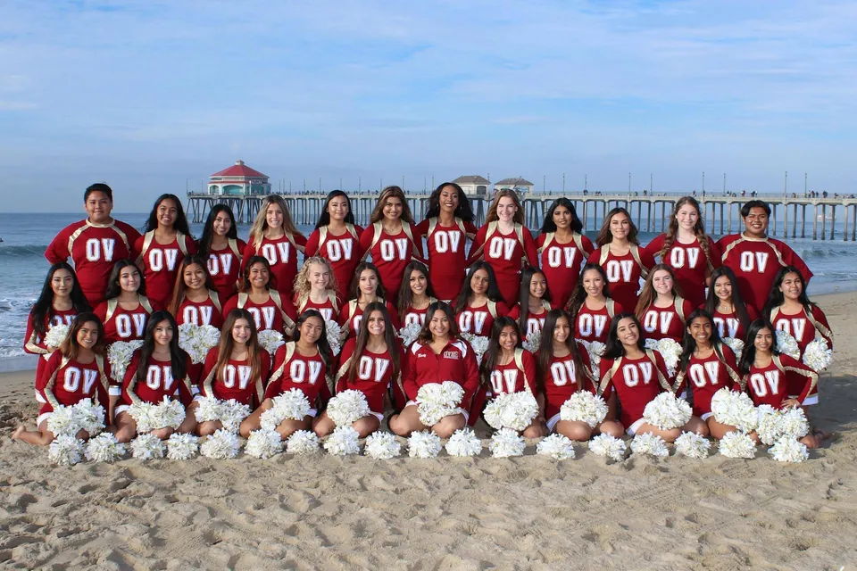 Un gruppo di cheerleader della Ocean View High School posa sulla spiaggia davanti a un molo e ad alcuni edifici sullo sfondo.