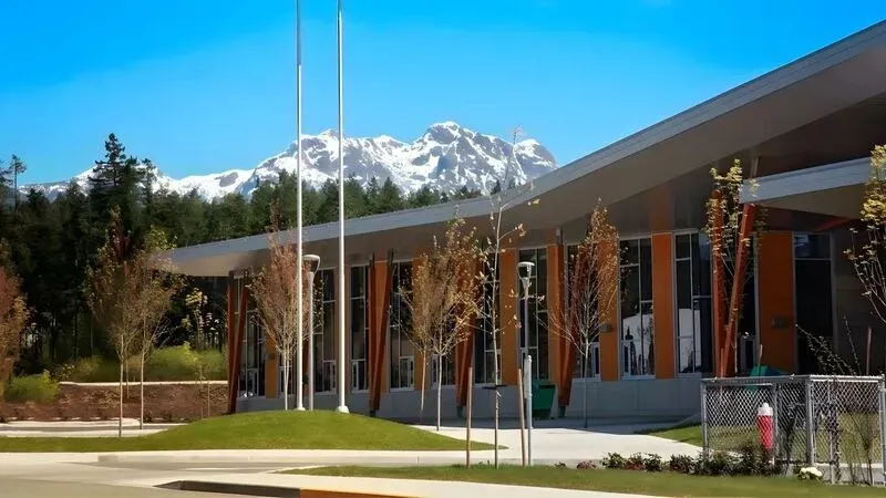 Edificio escolar bajo con tejado en voladizo y mástiles de bandera ante una cordillera nevada