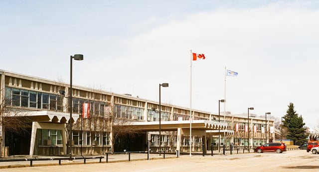 Das mehrstöckige Gebäude der Jasper Place High School mit einer Reihe von Flaggen im Vordergrund und einem Parkplatz sowie Bäumen im Hintergrund.