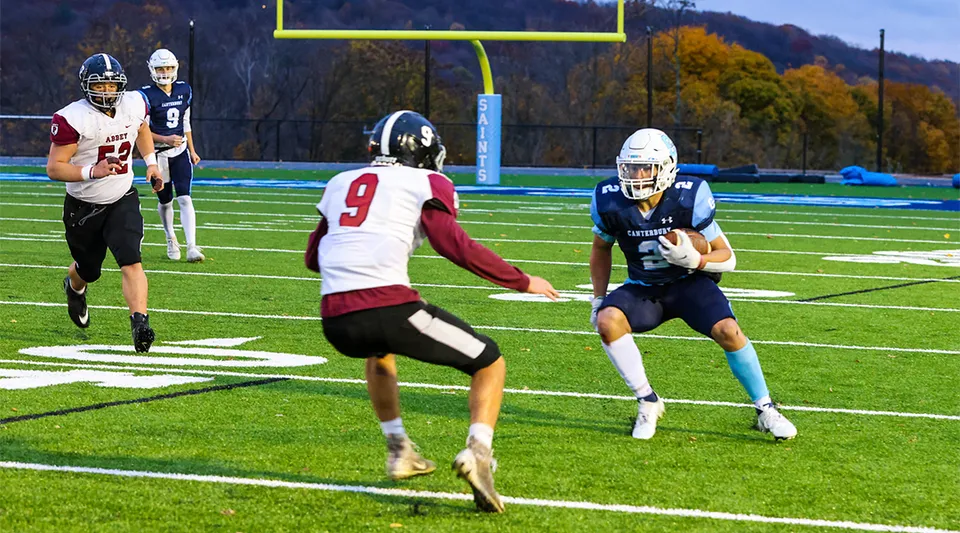 Due giocatori di football corrono sul campo da gioco della Canterbury School con foglie autunnali sullo sfondo.