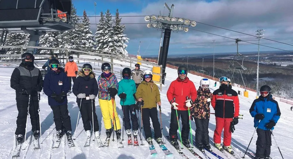 Un gruppo di studenti della Georgian Bay District Secondary School in colorata attrezzatura da sci si trova su un pendio innevato con skilift e montagne sullo sfondo.
