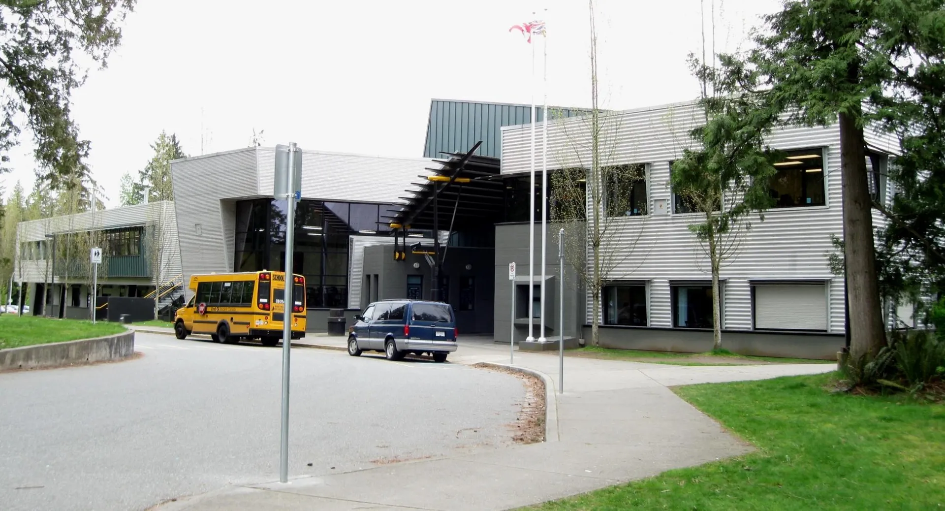 L'edificio moderno della Fraser Heights Secondary School con facciata in vetro e uno scuolabus giallo in primo piano è circondato da alberi e un'area verde.