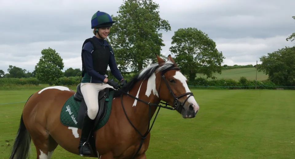 Una studentessa della Sibford School in abbigliamento da equitazione è a cavallo di un cavallo bianco e marrone su un prato con alberi sullo sfondo.