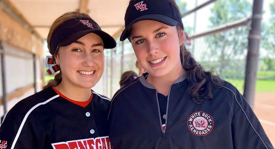 Due studentesse sorridenti della Thomas Haney Secondary School con cappellini da baseball e divisa sono in piedi davanti a un edificio.