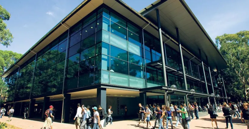 L'edificio moderno in vetro della Wollongong University con una folla di persone davanti, circondato da alberi e aree verdi.