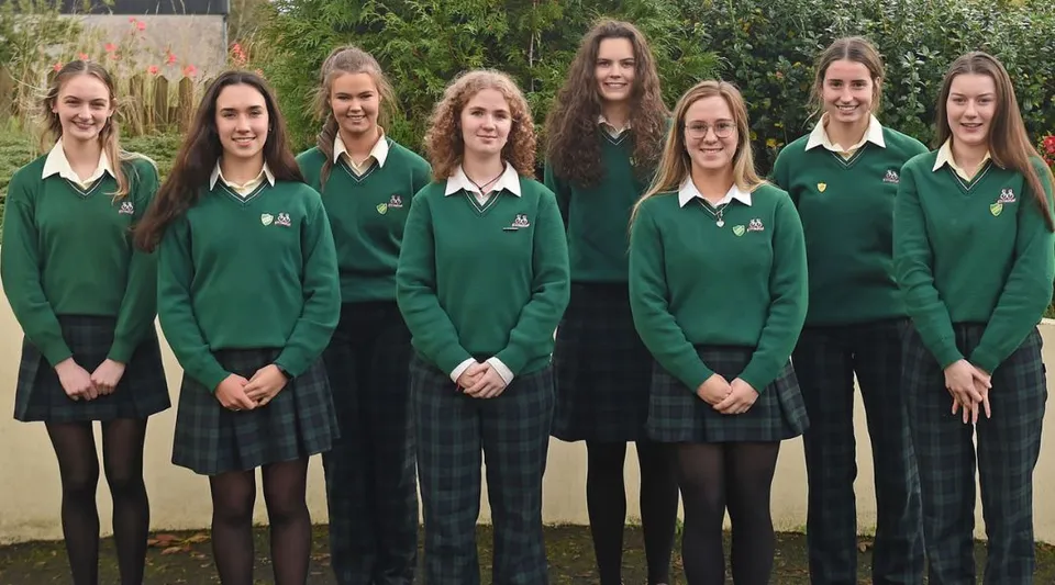 Un gruppo di studentesse della Sacred Heart School in uniforme scolastica verde si trova all'aperto su un prato davanti ad alberi e un edificio.