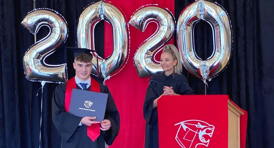Un diplomato della Burnaby Central Secondary School in toga e tocco tiene il suo diploma davanti a palloncini del 2020.
