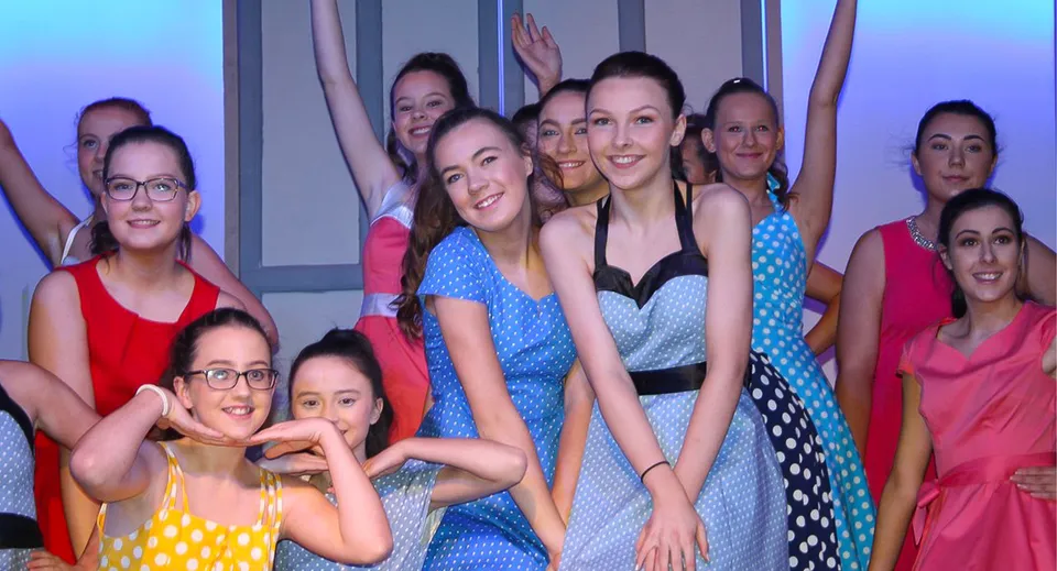 Un gruppo di donne vestite con abiti vivaci posa davanti a uno sfondo a motivi nel campus della Meán-Scoil Mhuire Gan Smál.
