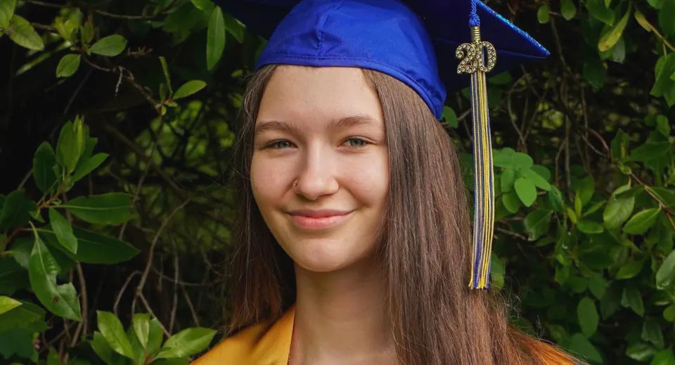 Una giovane donna con un cappello di laurea blu sorride all'aperto, tra il verde, nel campus delle Seattle Public Schools.