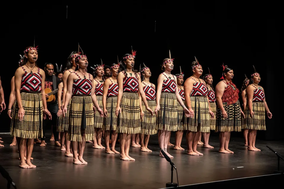 Un gruppo di studentesse della Gisborne Girls' High School si trova sul palco in costumi tradizionali d'ispirazione tribale con motivi elaborati.