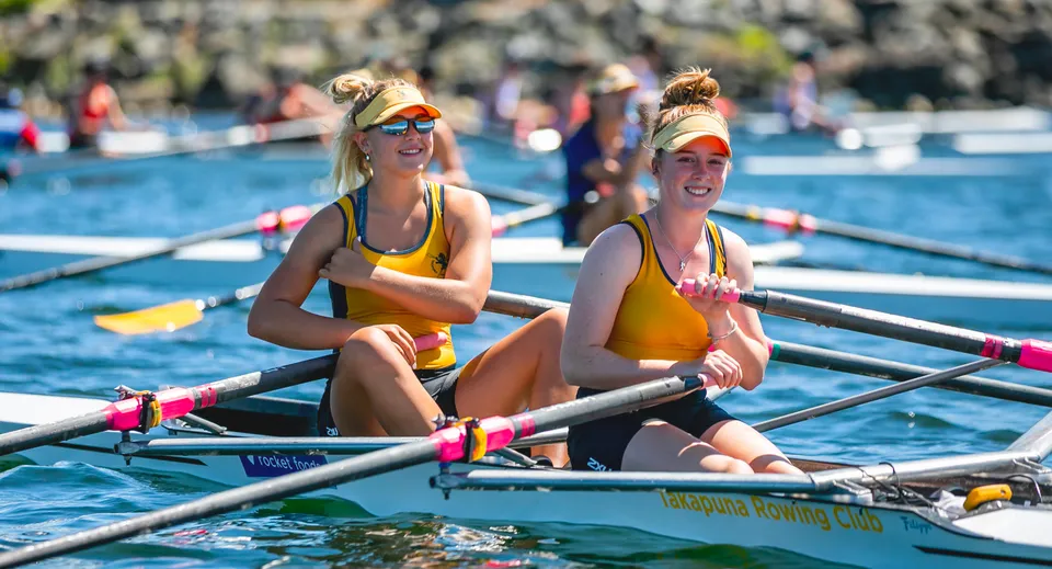 Due studentesse della Takapuna Grammar School siedono su una piccola barca a remi in acqua davanti agli spettatori.