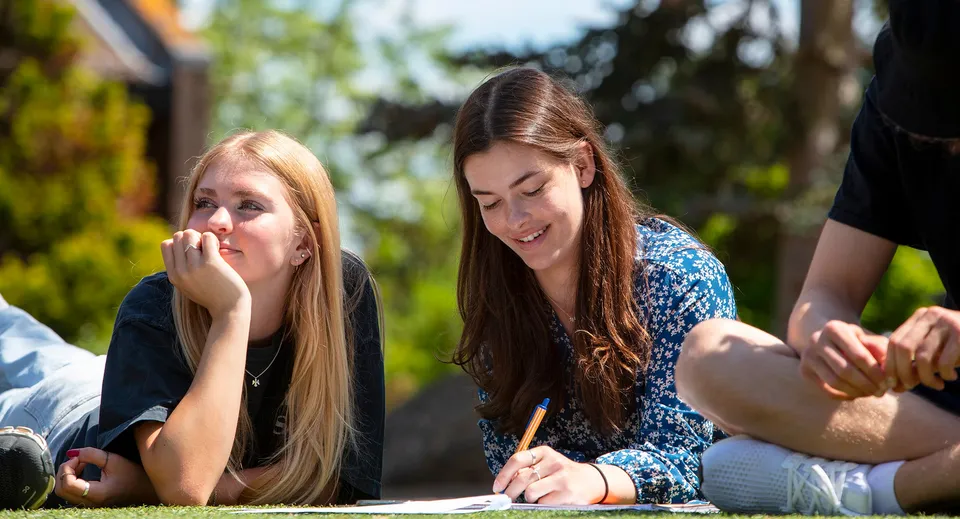 Due studentesse sedute sull'erba sotto gli alberi negli spazi esterni della ACS International School Cobham.