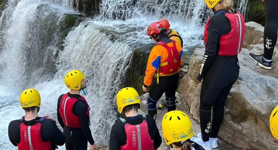 Un gruppo di studenti e studentesse della St. Clare’s School sta in piedi in abbigliamento protettivo colorato davanti a una cascata impetuosa.