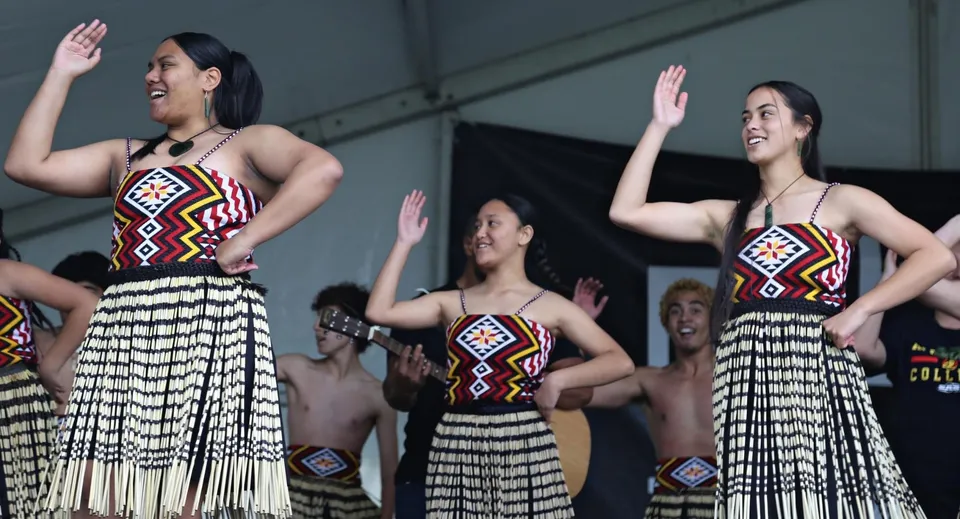 Tre donne eseguono una danza tradizionale sul palco davanti al pubblico dell'Alfriston College indossando abiti colorati.