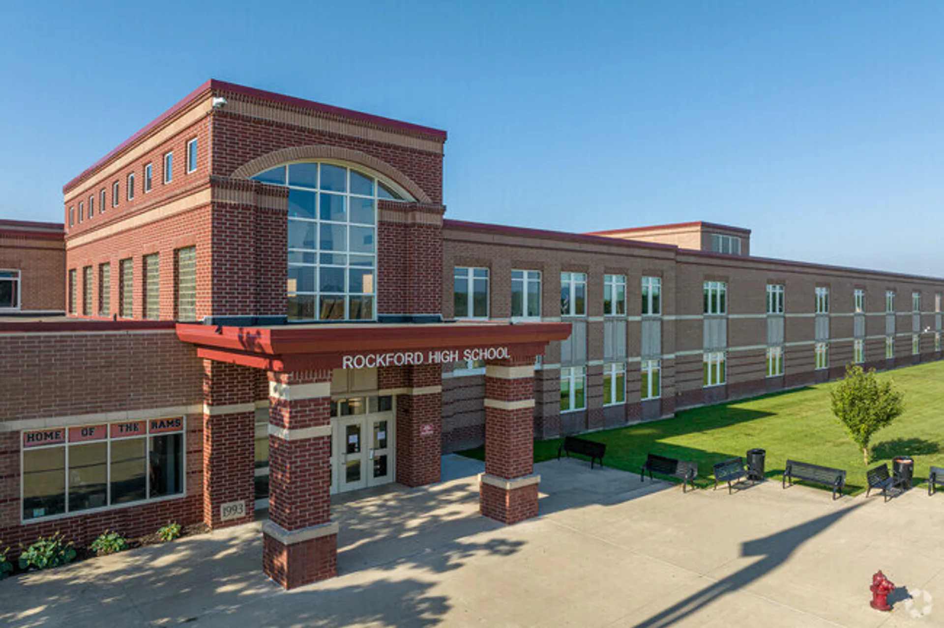 L'edificio a più piani in mattoni della Rockford High School, con finestre ad arco e area d'ingresso, sorge su un prato con panchine.
