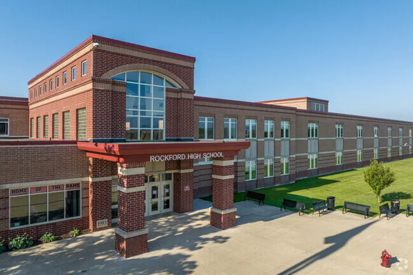 L'edificio a più piani in mattoni della Rockford High School, con finestre ad arco e area d'ingresso, sorge su un prato con panchine.