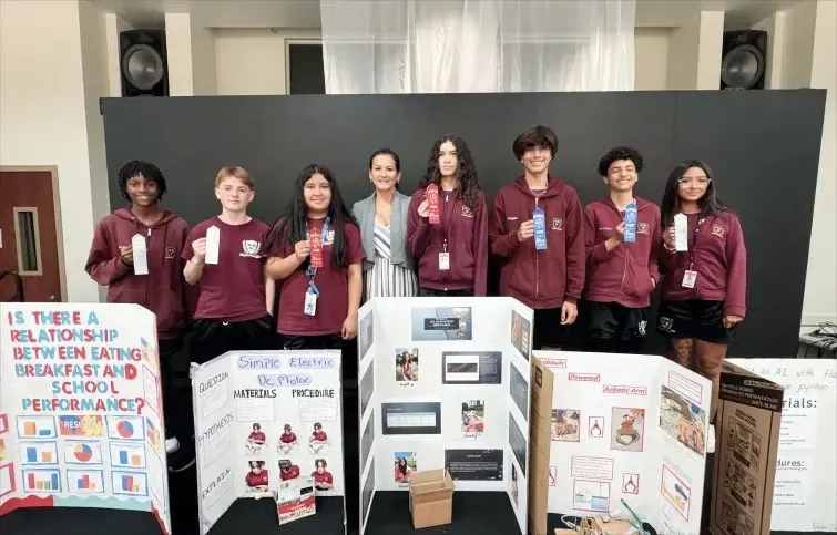 Estudiantes de la Elite Preparatory Academy presentan proyectos sobre trastornos alimentarios y rendimiento escolar frente a un panel informativo.