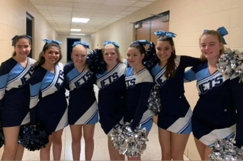 Siete animadoras con uniformes azules y pompones sonríen juntas en un pasillo escolar