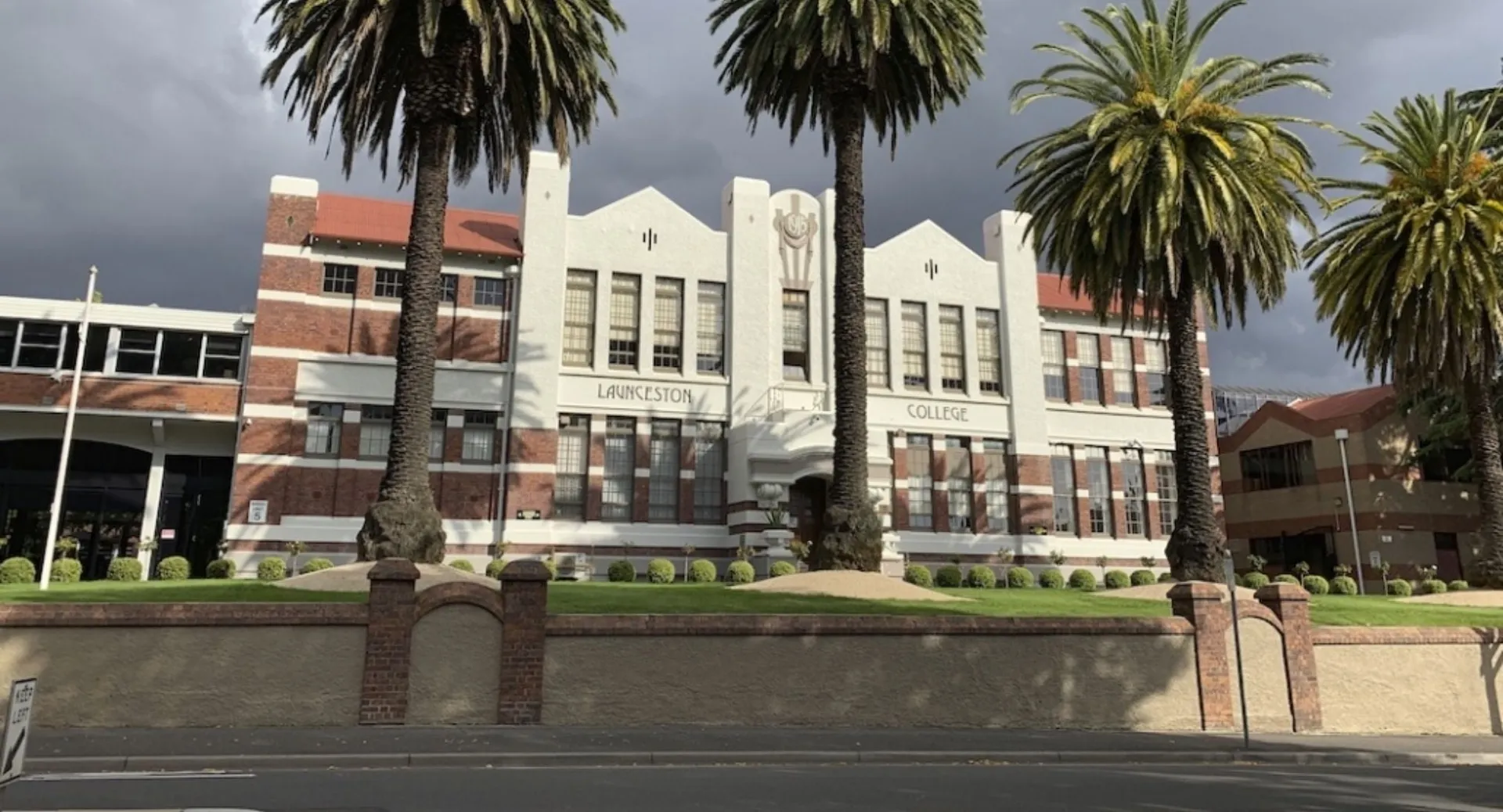 Il grande edificio decorato in mattoni del Launceston College con palme in primo piano sotto un cielo nuvoloso.