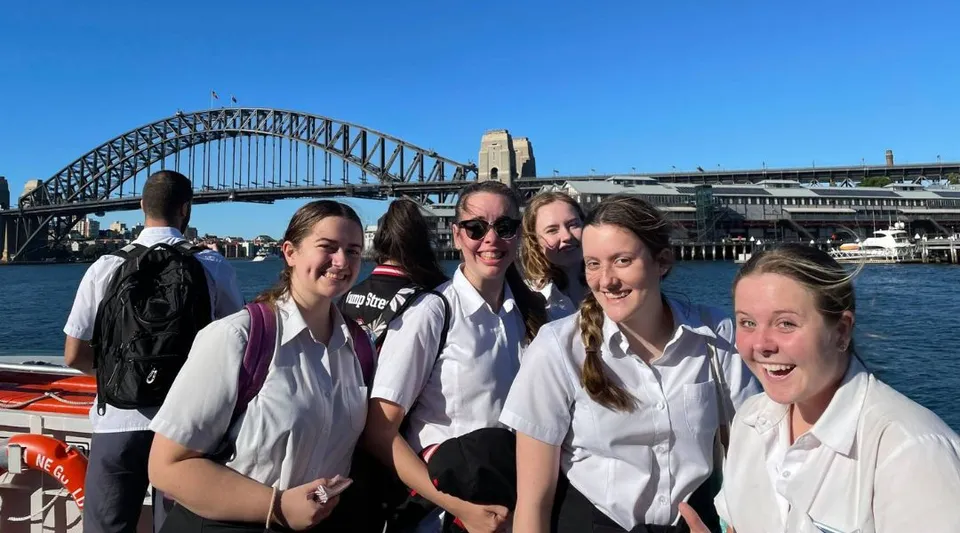 Un gruppo di giovani studentesse della Menai High School posa davanti al Sydney Harbour Bridge con lo skyline della città sullo sfondo.