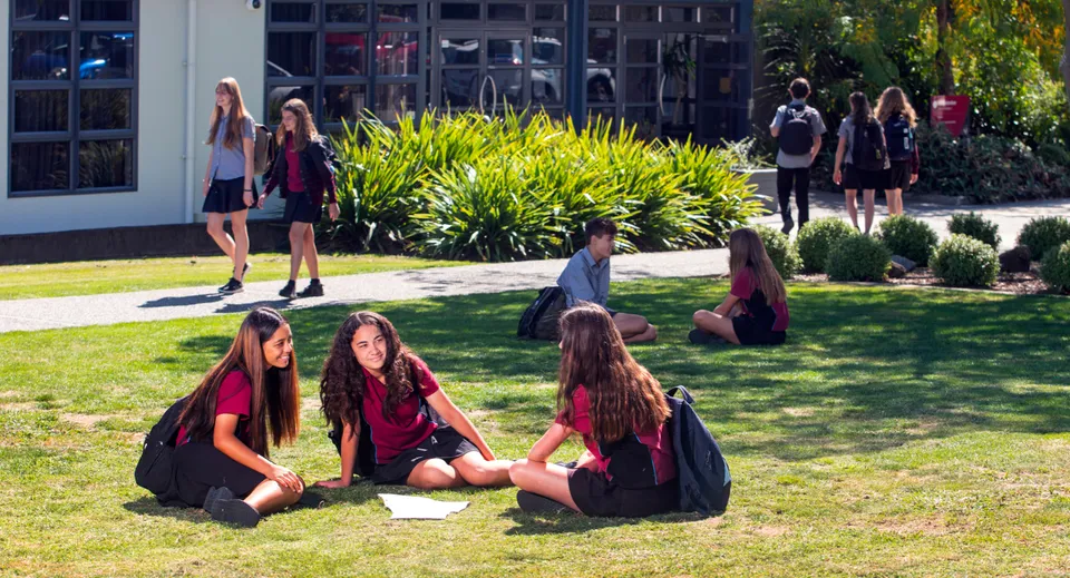 Un gruppo di giovani donne in toga nera siede sull'erba davanti a un edificio del Nayland College.