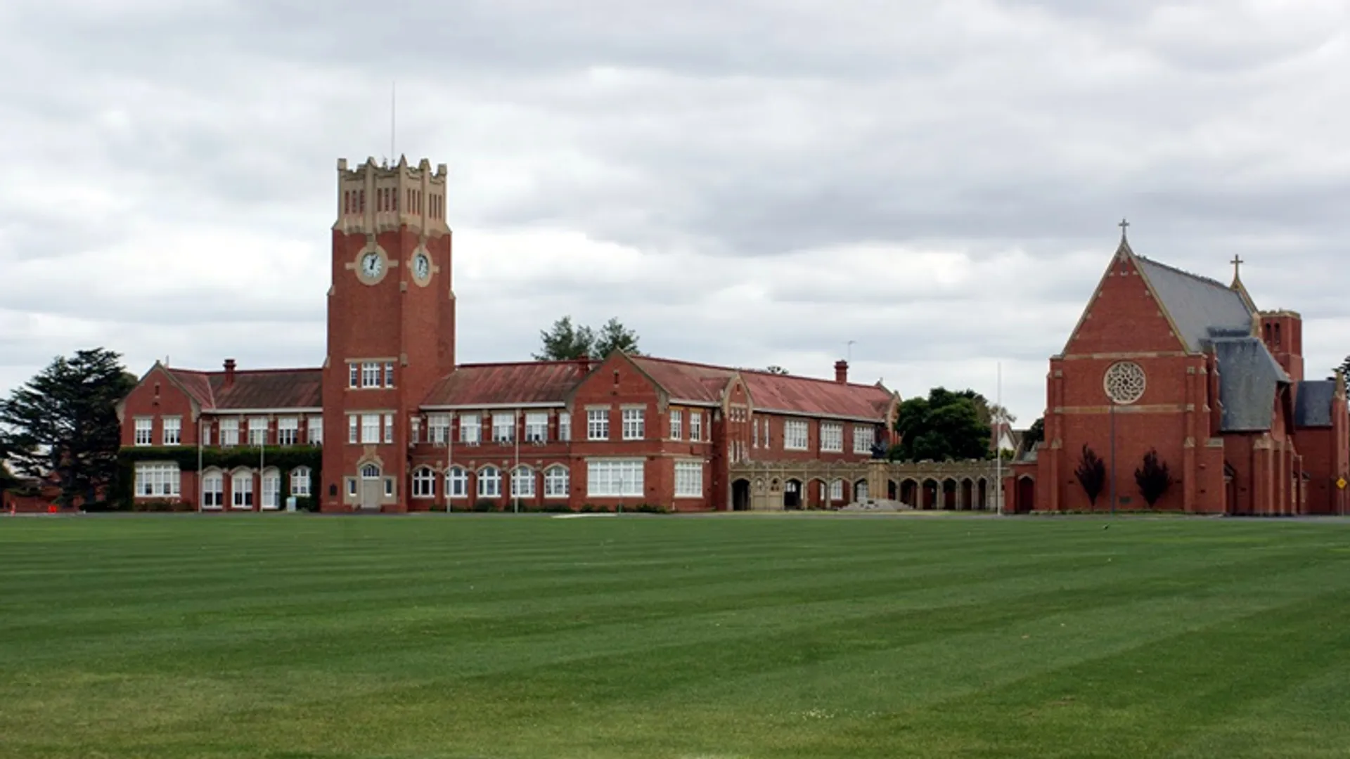 L'edificio in mattoni rossi con torre dell'orologio della Geelong Grammar School si erge su un prato verde sotto un cielo nuvoloso.