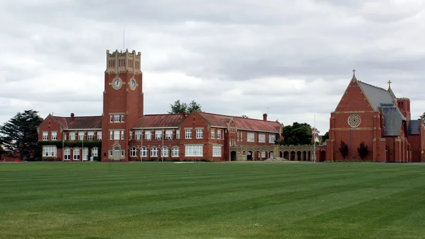 L'edificio in mattoni rossi con torre dell'orologio della Geelong Grammar School si erge su un prato verde sotto un cielo nuvoloso.