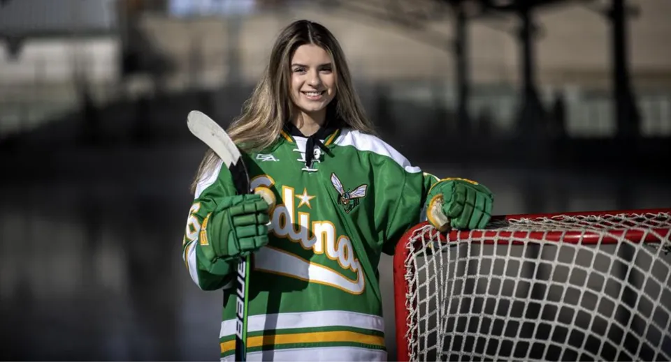 Una studentessa della Crowsnest Consolidated High School è in piedi con una mazza da hockey davanti a una porta da hockey.