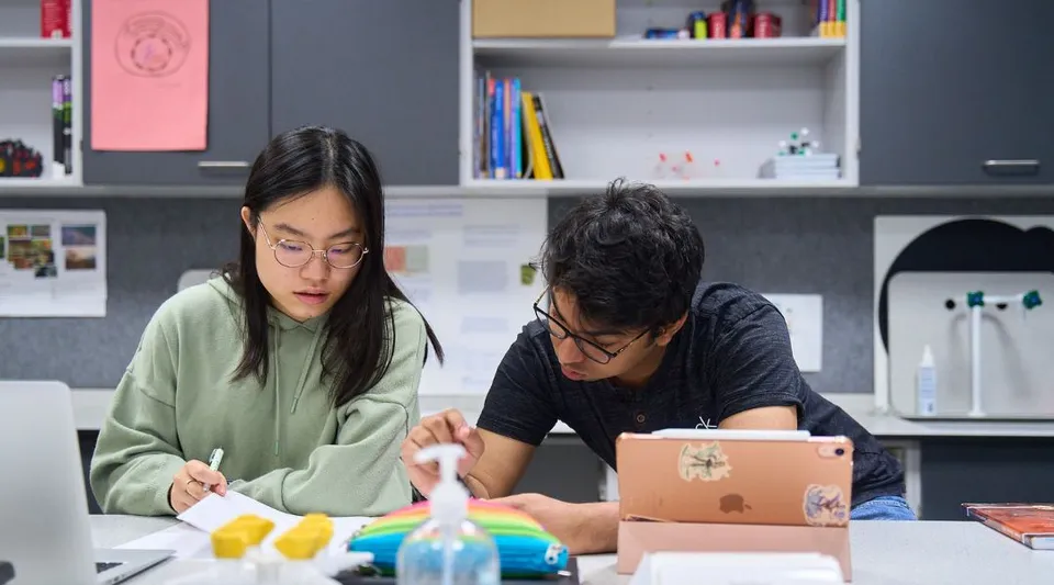 Due persone lavorano insieme a un progetto in una stanza con scaffali pieni di libri e materiale da ufficio nel campus della CATS Cambridge.