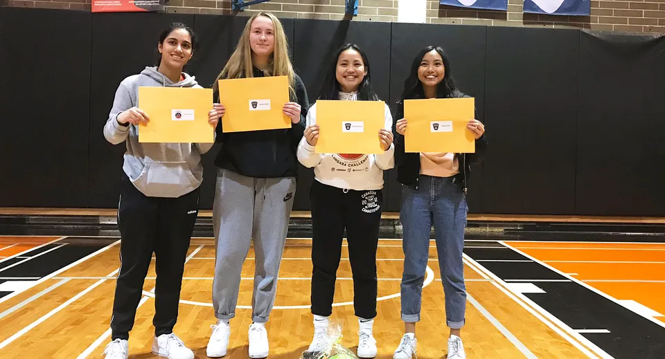 Quattro studentesse della John Oliver Secondary School sono in palestra e tengono cartelli gialli davanti al viso.