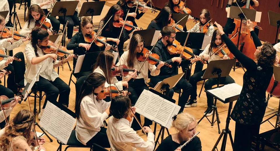 Un'ampia orchestra si esibisce sul palco della Jesus and Mary Secondary School in abito formale e suona vari strumenti a corda.