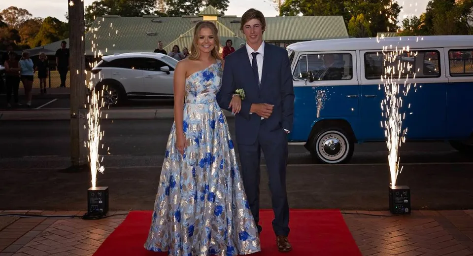 Una coppia in abito formale si trova sul tappeto rosso davanti a un vecchio van blu nel campus della Toowoomba Anglican School.