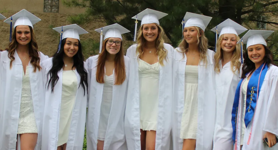 Un gruppo di giovani donne in tuniche bianche da diploma si trova insieme nel complesso del Coláiste Muire Crosshaven.