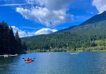 Dos jóvenes remando en un kayak por un lago rodeado de bosque y montañas