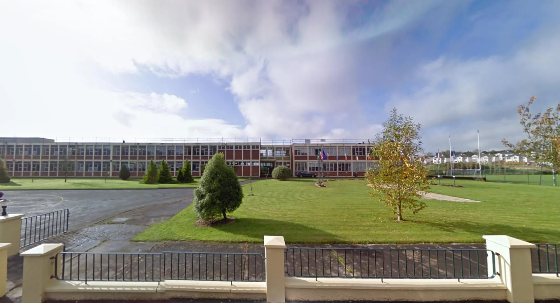 L'edificio scolastico della St. Aidan's Comprehensive School è visibile dietro una recinzione con un prato e alberi in primo piano sotto un cielo nuvoloso.
