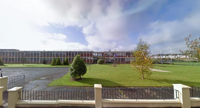 L'edificio scolastico della St. Aidan's Comprehensive School è visibile dietro una recinzione con un prato e alberi in primo piano sotto un cielo nuvoloso.