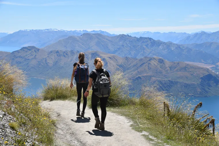 Zwei Wanderinnen mit Rucksäcken gehen auf einem Bergpfad mit Blick auf See und Gebirge