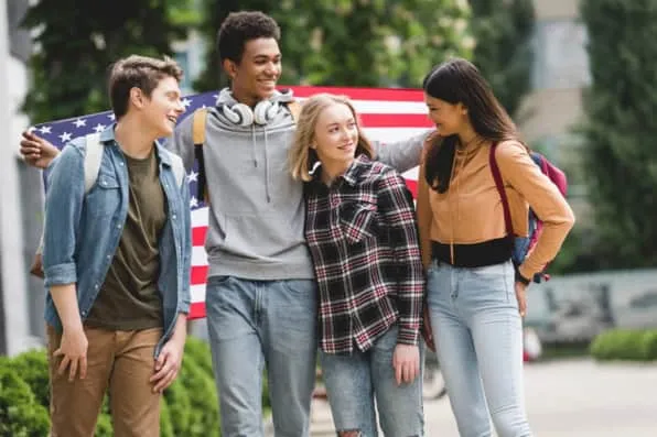 Cuatro jóvenes sonrientes con mochilas caminan juntos sujetando una bandera estadounidense al aire libre