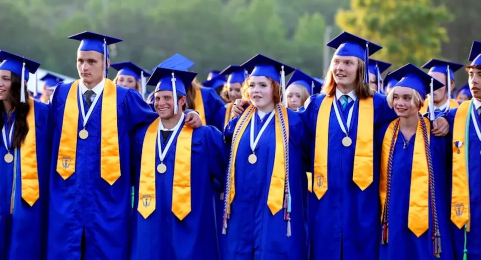 Un gruppo di diplomati in toga blu e oro è insieme davanti a un paesaggio naturale nel campus del Goderich District Collegiate Institute.