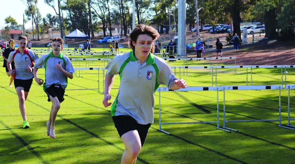 Una persona in abbigliamento sportivo bianco corre superando gli ostacoli su un campo in erba presso il campo sportivo della Kalamunda Senior High School.