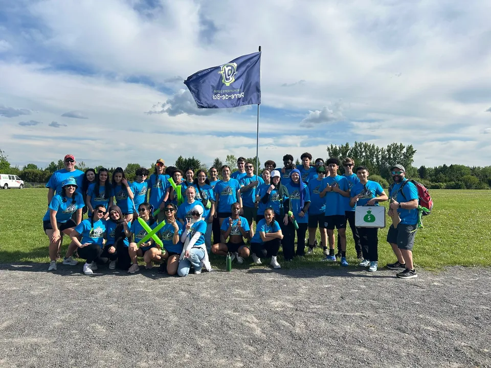 Un gruppo di studenti e studentesse in maglietta blu e verde sta davanti a una bandiera blu nel terreno della École secondaire publique Pierre de Blois.