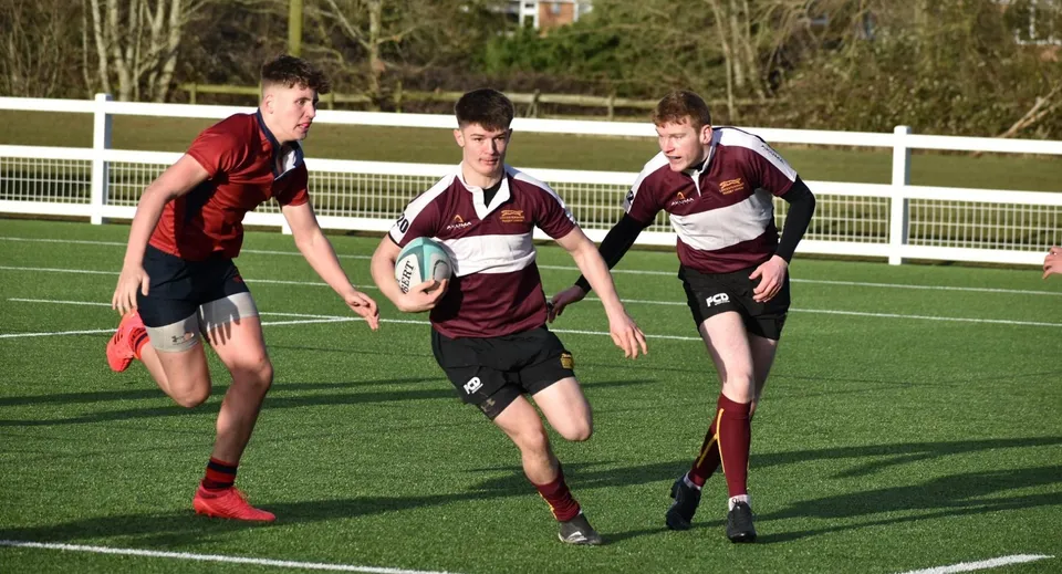 Tre studenti del Ratcliffe College giocano a rugby su un campo verde circondato da alberi.
