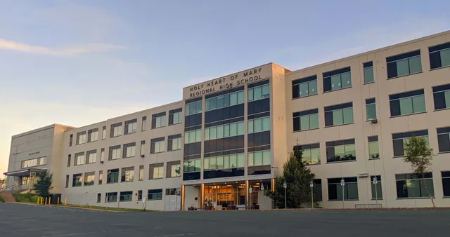 El edificio de varios pisos con ventanas de vidrio de la Holy Heart of Mary High School está rodeado de árboles y tiene un aparcamiento pavimentado en primer plano.