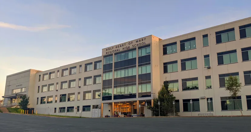 L'edificio a più piani con vetrate della Holy Heart of Mary High School è circondato da alberi e ha un parcheggio pavimentato in primo piano.