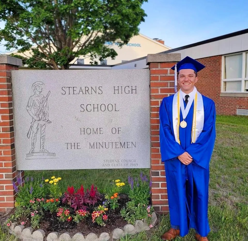 Un diplomato della Stearns High School in toga blu sta in piedi accanto all'insegna della scuola.