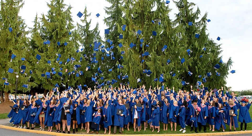 Un gruppo di diplomati in toga blu si trova davanti ad alti alberi sempreverdi nel campus della Brookswood Secondary School.