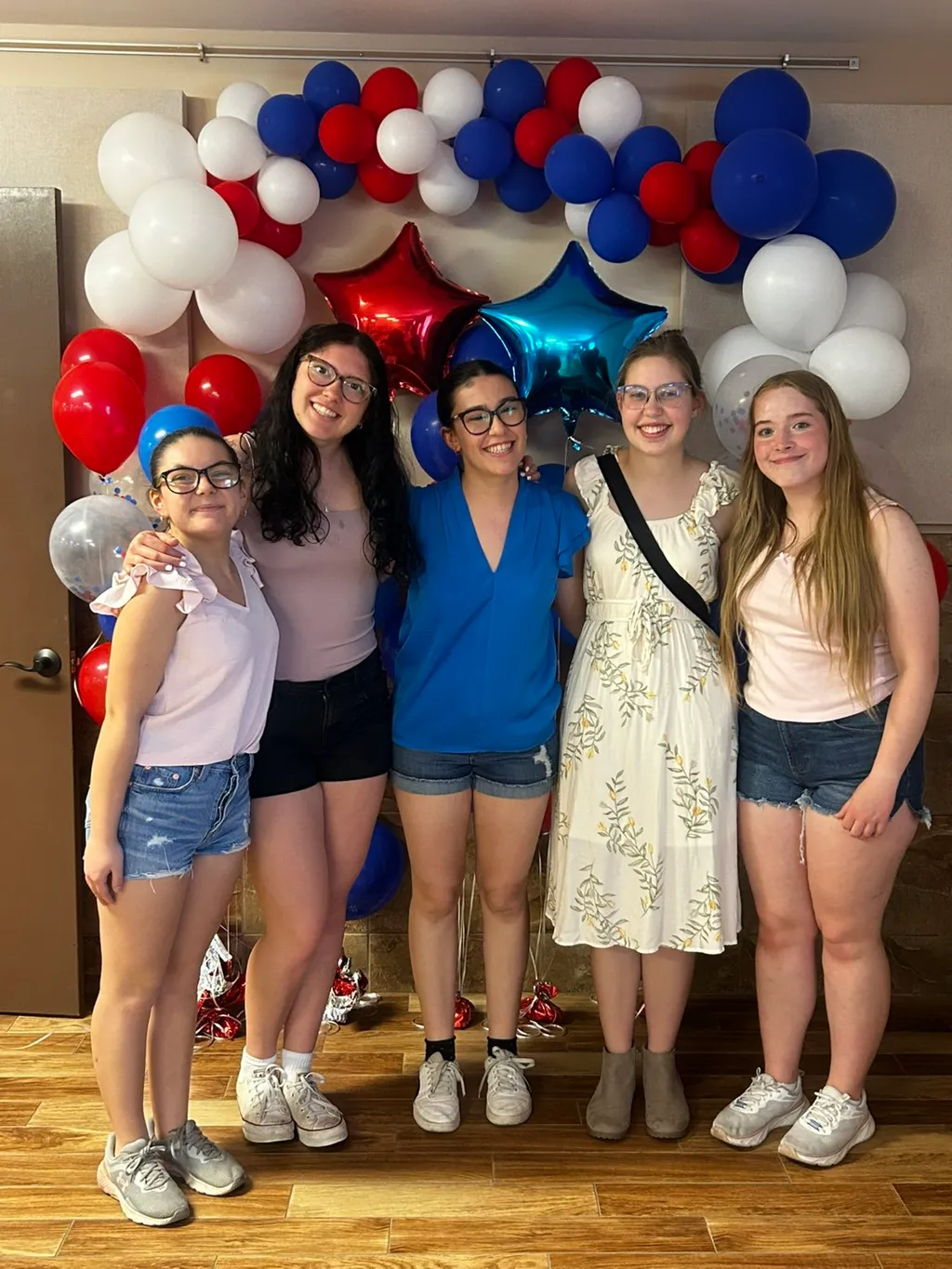 Cinque ragazze sorridenti davanti a un arco di palloncini rosso, bianco e blu con stelle