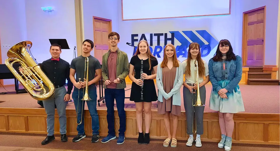 Un gruppo di studenti della Thunder Ridge High School si trova con strumenti musicali davanti a un grande banner Faith in una sala di una chiesa.
